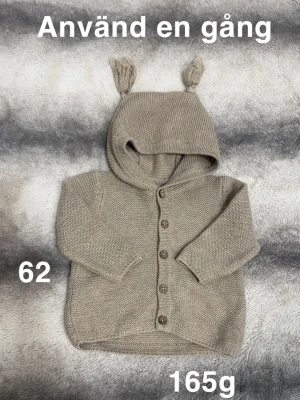 Brun/beige stickad kofta med huva och öron - Supersöt brun/beige stickad kofta med huva och små öron på toppen. Koftan har knappar framtill och långa ärmar, perfekt för kyliga dagar. Mysig och stilren design som passar till allt. 🌸 Använd en gång. Storlek 62