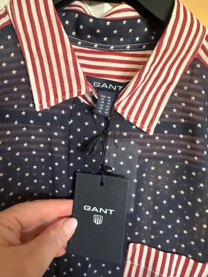 Blå skjorta med stjärnor och ränder från GANT - Säljer en unik skjorta från GANT i storlek 42 med blå botten och vita stjärnor, samt röda och vita ränder på krage, ärmar och bröstficka. Skjortan har knappar framtill och är långärmad. Materialet är tunn bomull, perfekt för dig som gillar att sticka ut. Helt ny och oanvänd 