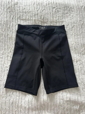 Svarta träningstights shorts från OTP - Säljer ett par svarta träningstights shorts från OTP i storlek XS. De har hög midja, praktiska sidofickor och är gjorda i ett stretchigt syntetmaterial som sitter tight och bekvämt. Perfekta för gymmet eller löprundan.