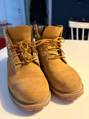 Timberland  - Klassiska Timberland boots i beige skinn med robust gummisula och gulbruna snören. Skorna har rund tå, snörning och svart vadderad kant upptill för extra komfort. Perfekta för höst och vinter, med slitstark konstruktion och ikonisk Timberland-logga på sidan. Innerfodret bak trasigt därav billigt pris. 
