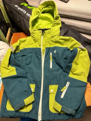 Grön skidjacka från Cross Sportswear - Snygg skidjacka från Cross. Jackan har huva, flera fickor med knappar och dragkedjor, samt vit dragkedja framtill. Liftkortsficka på armen. Materialet är slitstark polyester och jackan har långärmade ärmar. Perfekt för skidbacken. 