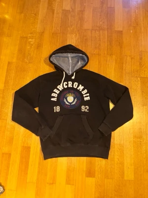 Svart Abercrombie & Fitch Vintage Hoodie - En supersnygg svart hoodie från Abercrombie and Fitch vintage-stil🤩Tröjan har ett tryck från Abercrombie med både text i tyg och dess logotyp👌En snygg och tjock tröja som denna passar perfekt nu under vintern❄️Tröjan är dessutom i mycket gott skick utan några defekter👍