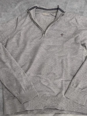 Grå half zip tröja från GANT - Snygg grå långärmad tröja från GANT med half zip och ribbade muddar. Klassisk ståkrage med svarta ränder och liten broderad logga på bröstet. Perfekt för dig som gillar stilren och enkel design. Passar runt 170-176.
