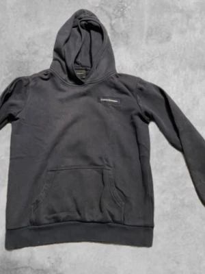 Mörkblå hoodie Peak Performance - Svart hoodie från Peak Performance i storlek 160. Klassisk modell med huva, magficka och diskret logga på bröstet. Mjuk insida och ribbade muddar vid ärmslut och nederkant. Perfekt för chill eller skola. Storlek 160.