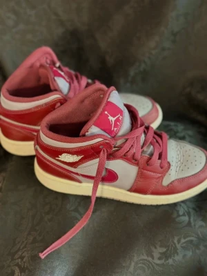 Nike Air Jordan 1 Mid röd och grå 35.5 - Nike Air Jordan 1 Mid sneakers i rött och ljusgrått med klassisk Jumpman-logga på plösen och Air Jordan Wings på sidan. Skorna har snörning, rund tå och platt sula. Ovandelen är i skinn och textil, med röda detaljer och Swoosh på sidan. Perfekt för dig som gillar streetstyle.