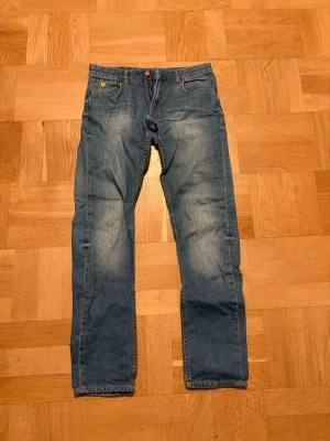 Jeans  - Klassiska  jeans i blå tvätt med raka ben och fem fickor. Jeansen har en knappgylf och tydliga slitningar för en cool, avslappnad look. Passar dig som gillar tidlös stil och vill ha ett par jeans som funkar till allt.