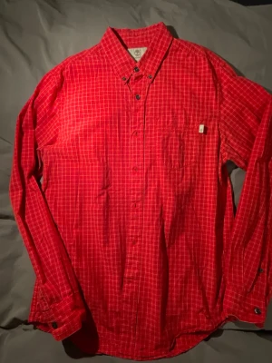 Rutig röd skjorta från Timberland - Säljer en röd rutig skjorta från Timberland i regular fit. Skjortan har klassisk krage, knappar framtill och en bröstficka med liten logga. Tillverkad i mjuk bomull och har långa ärmar. Perfekt för dig som gillar en avslappnad men snygg stil.
