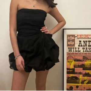 Svart axelbandslös kortklänning - Säljer en svart axelbandslös kortklänning med voluminös kjol och draperad överdel. Klänningen har en synlig dragkedja bak och är tillverkad i ett tunt, transparent tyg som ger en cool och festlig look. Perfekt för dig som vill sticka ut med en unik siluett.