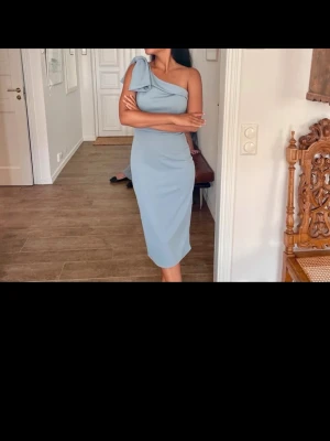 Ljusblå oneshoulder klänning - Säljer en elegant ljusblå klänning med oneshoulder-design och en snygg rosettdetalj på axeln. Klänningen är figurnära och har en längre, smal siluett som ger en stilren look. Perfekt för dig som vill sticka ut med en modern och chic stil.