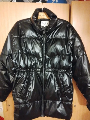 Svart pufferjacka från NA-KD - Säljer en svart, glansig pufferjacka från NA-KD i storlek 36. Jackan har hög krage, dragkedja framtill och två stora fickor. Midjan har snörning för justerbar passform och jackan är perfekt för kalla dagar. Materialet är syntet och jackan är långärmad.