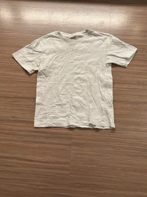 Vit t-shirt från Kappahl - En enkel vit t-shirt från Kappahl i storlek 134/140. Modellen har rund hals och korta ärmar, tillverkad i mjuk bomull som är skön mot huden. Perfekt för dig som gillar basic och stilrena plagg. Passar till många olika outfits.