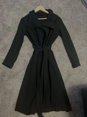 Svart lång kappa med bälte - Säljer en svart lång kappa med bred krage och knytskärp i midjan. Jackan har långa ärmar och två fickor framtill. Perfekt för dig som vill ha en stilren och enkel look under höst och vinter.