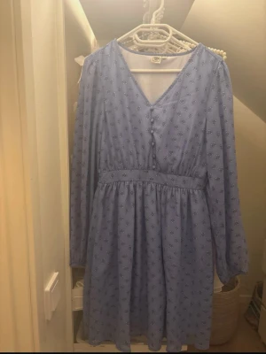 Blommig blå klänning  - Söt blå klänning från Ullared med små blommor, v-ringad hals och knappar framtill. Klänningen har långärmade ärmar med resår vid mudden och ett markerat midjeband. Perfekt för dig som gillar en feminin och avslappnad stil. Jag har aldrig använt klänningen.