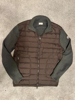 Grå Moncler cardigan  - Säljer en brun och mörkgrå kofta från Moncler med quiltade dunpaneler framtill och stickade ärmar. Tröjan har dragkedja hela vägen, hög krage och två fickor med dragkedja. Perfekt för kyliga dagar och riktigt snygg till jeans. Stolek xl passar L