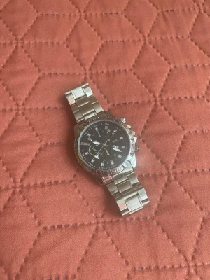 Klocka - Bilderna visar en silvrig armbandsklocka från SOKI med svart urtavla och metallarmband som inte går att justera.