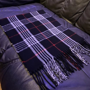 Burberry Halsduk - Säljer en Burberry halsduk med klassiskt rutigt mönster i vitt och röda linjer. Halsduken har snygga fransar i ändarna och är gjord i ett mjukt material som känns skönt mot huden. Perfekt att slänga på sig när det är kyligt ute.