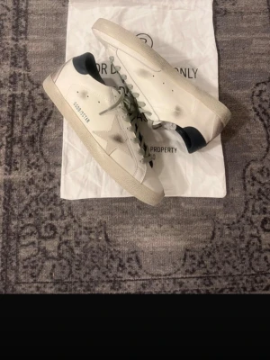 Golden Goose vita sneakers med stjärna - Säljer ett par vita Golden Goose sneakers med den ikoniska stjärnan på sidan och grå snörning. Skorna har svarta detaljer vid hälen och en vintage-inspirerad look med slitningar på sula och tå. Tillverkade i skinn och levereras med dustbag. Kolla dom andra skorna också kan ge bra pris för båda två!