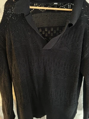 Svart stickad tröja från Bik Bok - Säljer en svart stickad tröja från Bik Bok med krage och v-ringning. Tröjan har ett genombrutet mönster och långa ärmar. Perfekt för lager på lager och ger en chill men ändå snygg look.