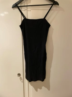 Svart tight kortklänning med smala axelband - En stilren svart kortklänning med smala axelband och rak halsringning. Klänningen har en tight passform och är perfekt för dig som gillar en enkel men snygg look. Materialet känns mjukt och följsamt mot kroppen.