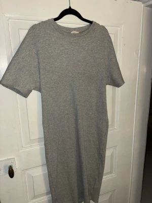 Grå ribbad t-shirtklänning H&M - Enkel och stilren grå t-shirtklänning från H&M i ribbad bomull. Klänningen har korta ärmar, rund halsringning och rak passform. Perfekt för en chill och avslappnad look.
