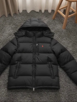 Svart jacka från Polo Ralph Lauren - Säljer en svart dunjacka från Polo Ralph Lauren med röd broderad logga på bröstet. Jackan har huva, dragkedja framtill och två sidofickor. Puffig modell som är perfekt för kalla dagar. Klassisk och stilren design med RL-detalj på dragkedjan.