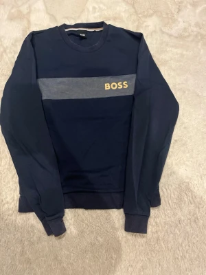 Mörkblå sweatshirt från Hugo BOSS - Säljer en mörkblå sweatshirt från Hugo BOSS med grått band över bröstet och gul BOSS-logga. Tröjan har rund hals, långa ärmar och ribbade muddar. Tillverkad i mjuk bomull, perfekt för en clean och stilren look.