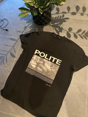 Svart t-shirt Young Poets Society - Svart t-shirt från Young Poets Society med tryck på framsidan där det står 'POLITE' och en grafisk bild under. Klassisk rund hals och korta ärmar. Materialet är mjuk bomull som känns skönt mot huden. Perfekt för dig som gillar streetwear och tydliga prints.