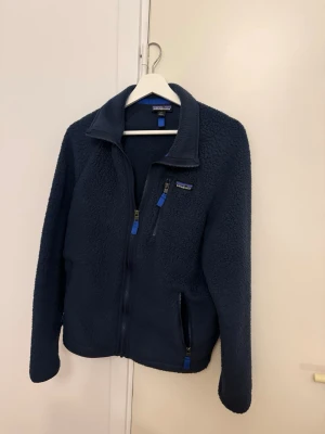 Mörkblå fleecejacka från Patagonia - Mysig mörkblå fleecejacka från Patagonia med hel dragkedja och tre fickor med blå detaljer. Jackan har hög krage och långärmade ärmar, perfekt för kyliga dagar. Snygg och enkel att matcha med jeans eller träningskläder.