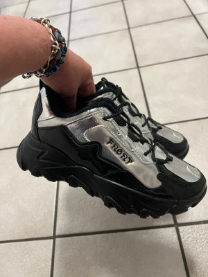 Chunky silvriga sneakers från Frory - Säljer ett par chunky sneakers från Frory i metallic silver och svart. Skorna har en rejäl sula, snörning och detaljer i syntetmaterial. Insidan är fodrad med svart fuskpäls för extra komfort. Perfekta för dig som gillar statement-skor med futuristisk vibe.