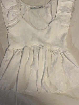 Vit blus med volangdetaljer XS - Säljer en söt vit blus från 157 i storlek XS. Blusen har volangdetaljer vid axlarna och en rundad halsringning. Materialet är lätt och har en diskret struktur som ger en fräsch känsla. Perfekt för ljusa dagar och enkel att matcha med jeans eller kjol.