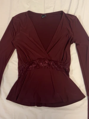 Vinröd omlottopp från Gina Tricot - Säljer en vinröd omlottopp från Gina Tricot i storlek XXS. Toppen har lång ärm, v-ringad hals och ett snyggt spetsband med blommönster i midjan. Materialet är mjukt och stretchigt, perfekt för att skapa en feminin siluett.