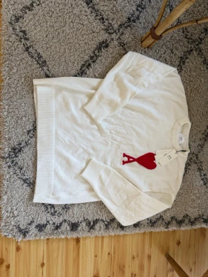 Vit stickad tröja från Ami Paris - Säljer en vit stickad tröja från Ami Paris med ett stort rött hjärta och bokstaven A på bröstet. Tröjan har långa ärmar, ribbade muddar och rund hals. Materialet är mjuk ull och designen är clean med en snygg detalj framtill. Perfekt för dig som gillar stilrena plagg med en twist.