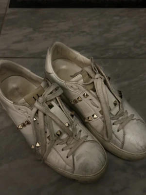 Valentino Rockstud vita sneakers - Säljer ett par vita Valentino Rockstud sneakers med coola guldfärgade nitar och snörning. Skorna är tillverkade i skinn och har en platt sula med rund tå. Märket syns både på insidan och under sulan. Perfekt för dig som gillar lyxiga detaljer. Passar mig som har 38