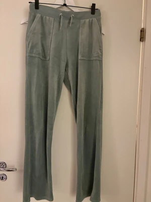 Ljusgröna Juicy Couture byxor - Säljer ett par ljusgröna mjukisbyxor från Juicy Couture i mjuk sammet med breda fickor fram och bak, samt snörning i midjan. Byxorna har en relaxed passform och raka ben, perfekt för chill dagar. Ikonisk JC-logga broderad på ena fickan. OBS! Storlek 14/15 - kidsbrandstore 