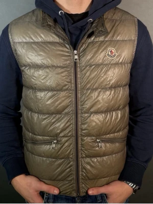 Beige dunväst från Moncler - Säljer en beige dunväst från Moncler med quiltad design och glansig finish. Västen har två fickor med dragkedja framtill och klassisk Moncler-logga på bröstet. Perfekt att slänga över en hoodie för en chill look.