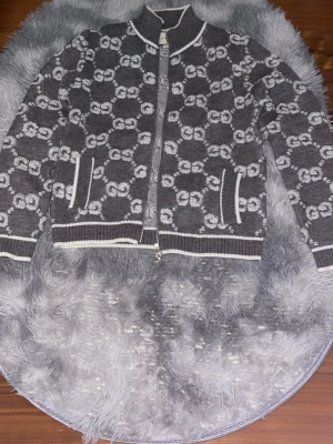 Grå kofta från Gucci med mönster - Säljer en snygg grå kofta från Gucci i 100% ull med vit GG-mönster över hela plagget. Koftan har dragkedja framtill, ribbade muddar och vita detaljer vid krage, fickor och nederkant. Perfekt för dig som gillar lyxiga märken och stilrena mönster.