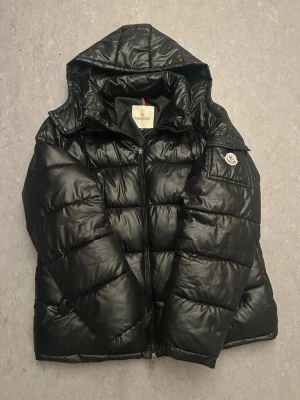 Svart Moncler pufferjacka med huva - Säljer en svart pufferjacka från Moncler med huva och klassisk logga på ärmen. Jackan har quiltad design, dragkedja framtill och innerfoder med serietidningsmönster. Perfekt för kalla dagar och riktigt snygg streetstil. Loggan är bra men den är lite limat och är det lite problem med dragkedjan men kommer fixa den så de bårde inte inte va några problem.