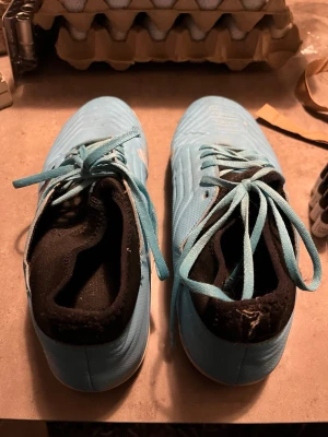 Adidas ljusblå fotbollsskor med snörning - Säljer ett par ljusblå fotbollsskor från Adidas med vit sula och svart insida. Skorna har snörning och är tillverkade i syntetmaterial. Snyggt mönstrad ovansida och Adidas-logga på sulan. Perfekta för inomhusspel och träning.