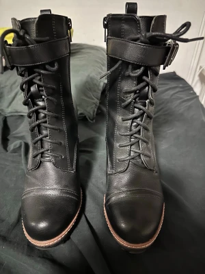 Svarta snörade stövletter med klack - Säljer ett par svarta stövletter i skinnimitation med grov klack, snörning framtill och spänne upptill. Dragkedja på sidan för enkel påtagning.      Använda en gång litet märke som knappt syns på ena skon som är med på bild 4. 