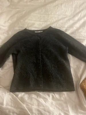 Svart kofta från 157 med knappar - Mysig svart kofta från 157 med lång ärm och knäppning framtill. Koftan är gjord i ett fluffigt, stickat material som känns både varm och stilren. Perfekt för lager på lager och enkel att matcha till olika outfits.den är använd 1-2 gånger.