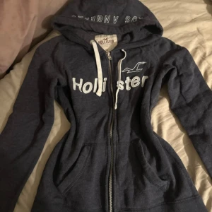 Vintage Mörkblå/grå hoodie från Hollister XS - Mörkblå hoodie från Hollister med vit logga och text framtill. Tröjan har huva med vita snören, dragkedja hela vägen och två fickor framtill. Perfekt för en chill och avslappnad stil. Lite nopprig men det är en självklarhet då den är vintage. 