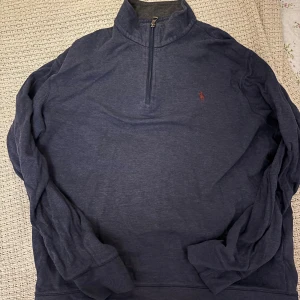Blå half zip tröja från Polo Ralph Lauren - Mörkblå långärmad tröja från Polo Ralph Lauren med klassisk broderad logga på bröstet. Tröjan har half zip-dragkedja och hög krage, perfekt för lager-på-lager. Tillverkad i mjuk bomullsblandning för en skön känsla.