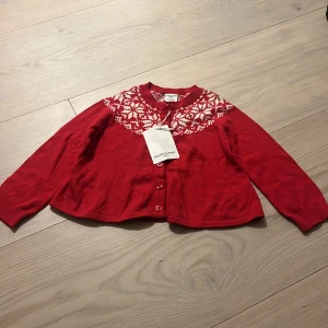 Röd kofta med mönster från Polarn O. Pyret - Röd kofta från Polarn O. Pyret med klassiskt vitt mönster över axlarna. Rund hals, knäppning framtill med röda knappar och långa ärmar. Perfekt för vintern och julen, stickad i mjukt material.