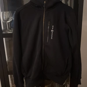 Svart zip hoodie från Sail Racing - Svart zip hoddie från Sail Racing med huva och hel dragkedja framtill. Hoodien har en bröstficka med vertikal dragkedja och logotyptryck, samt två sidofickor. Materialet är troligtvis polyester och passformen är normal. 