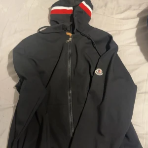 Svart zip hoodie  från Moncler - Svart zip hoodie  från Moncler med huva och dragkedja framtill. Tröjan har Moncler-logga på ärmen och snygga rödvita ränder på huvan. Perfekt för dig som gillar stilrena och sportiga plagg med exklusiv känsla. Storlek XL men är större i storleken hade passat vikterna mellan 80kg - 100 kg 