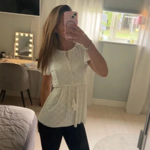 Vit blus med spets och knappar - Supersöt vit blus med broderade spetsdetaljer. Blusen har korta, volangiga ärmar och ett knytband i kragen. Perfekt för dig som gillar romantisk och feminin stil.