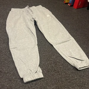 Grå mjukisbyxor från Nike - Snygga grå mjukisbyxor från Nike med dragsko i midjan och resår vid bensluten. Byxorna har en klassisk Nike-logga på låret och är tillverkade i mjukt bomullsmaterial för maximal komfort. Perfekta för chill eller träning.