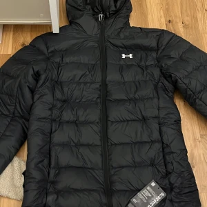 Svart pufferjacka från Under Armour - Säljer en svart pufferjacka från Under Armour med huva och dragkedja framtill. Jackan har quiltad design och är perfekt för kyliga dagar. Klassisk logga på bröstet och en clean look som funkar till det mesta.