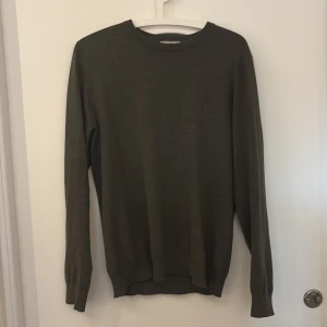 Mörkgrön stickad tröja från Bläck - En stilren mörkgrå stickad tröja från Bläck i storlek S. Tröjan har rund halsringning, ribbade muddar vid ärmslut och nederkant samt långärmad passform. Perfekt för dig som gillar enkel och clean look.