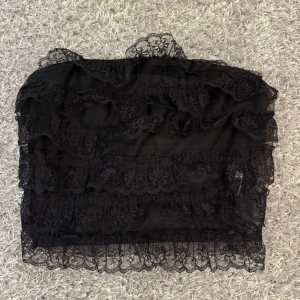 Svart bandeau tubtopp med spets - Säljer en svart bandeau tubtopp med flera lager spets och volanger. Toppen är axelbandslös och har en transparent, romantisk look med fina spetsdetaljer och croppad passform. Perfekt att styla med jeans eller kjol för en cool vibe.